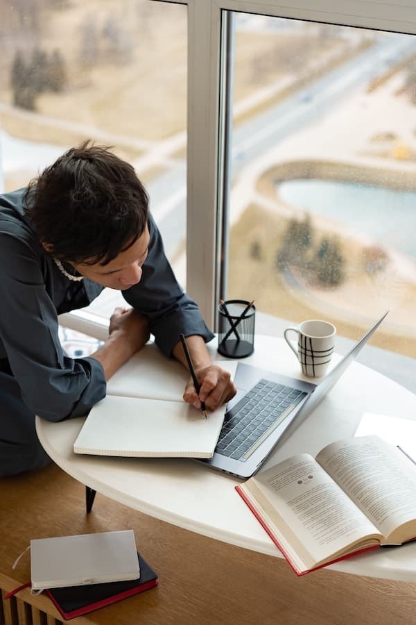 Personne écrivant dans un carnet entre un ordinateur portable et plusieurs livres ouverts