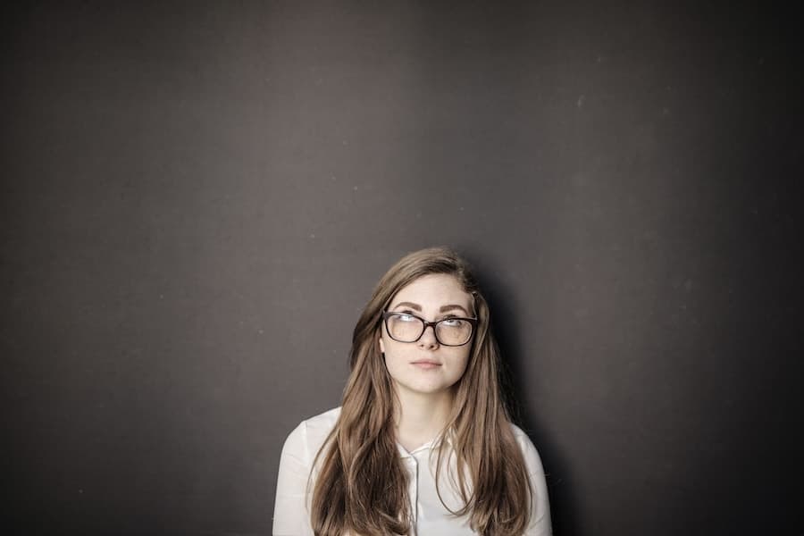 Jeune femme portant des lunettes, assise devant un fond sombre et regardant vers le haut