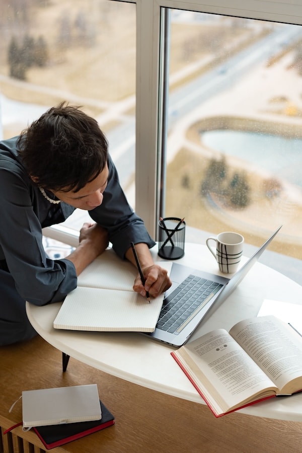 Personne écrivant dans un carnet entre un ordinateur portable et plusieurs livres ouverts